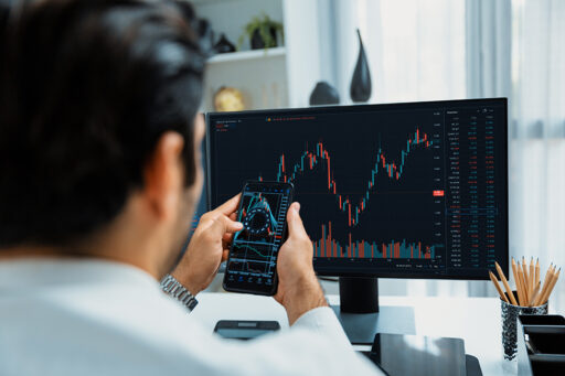 Mann analysiert Börsencharts auf Smartphone und Monitor, Darstellung von Aktienhandel und Finanzanalyse in Echtzeit.